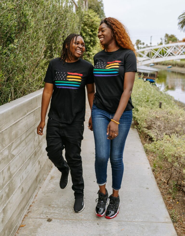 two Black women walking. They are both wearing t-shirts that have a large US flag across the chest; the flag's stripes are rainbow colored like the Pride flag.